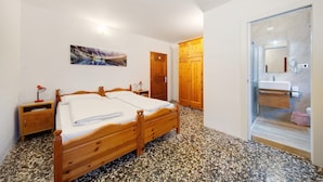 Quarto com cama de madeira, mesa de cabeceira, luminária na parede, porta e banheiro com pia e espelho.
