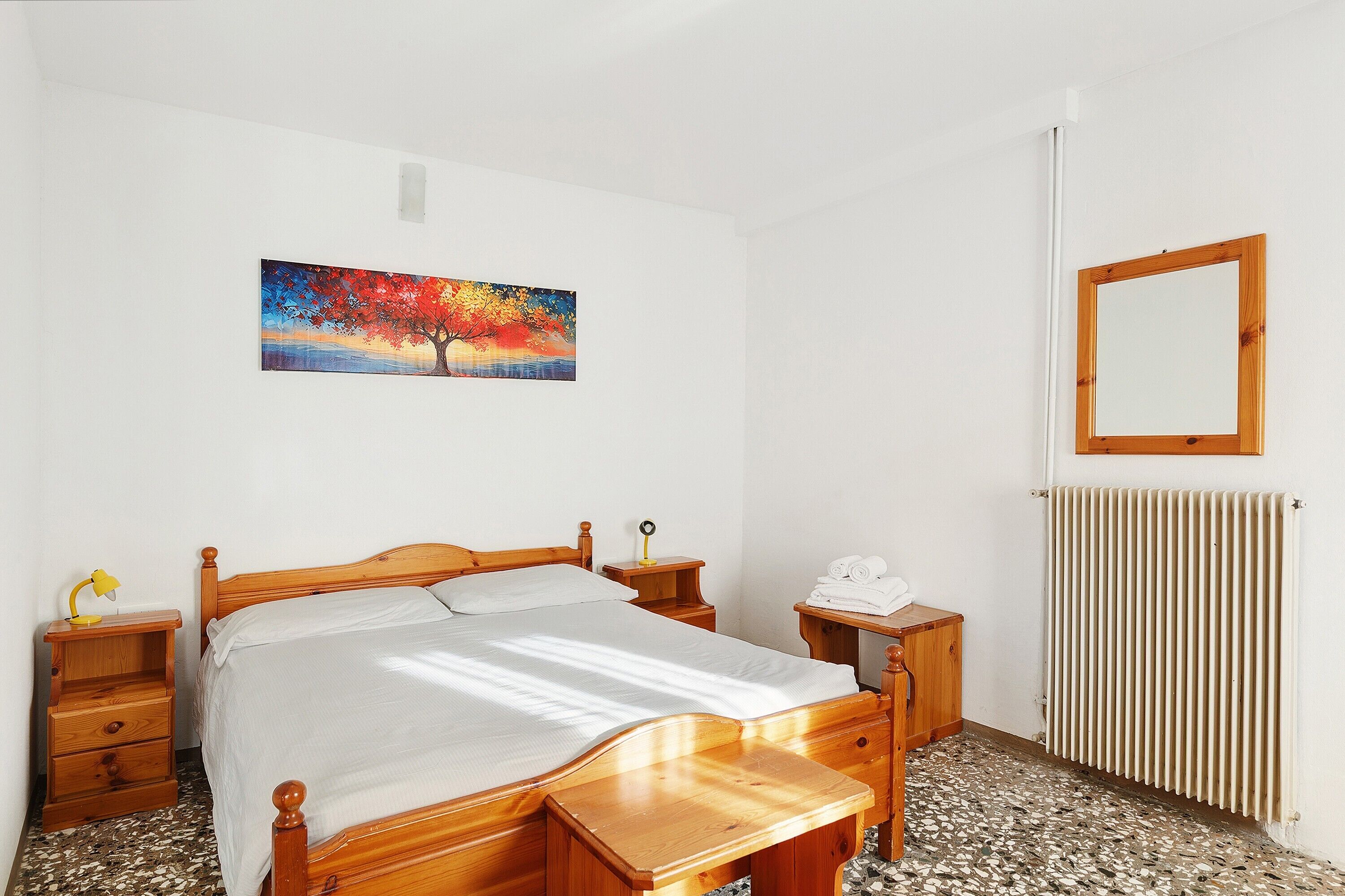 A bedroom with a wooden bed, a nightstand with a lamp, a painting on the wall, and a radiator.