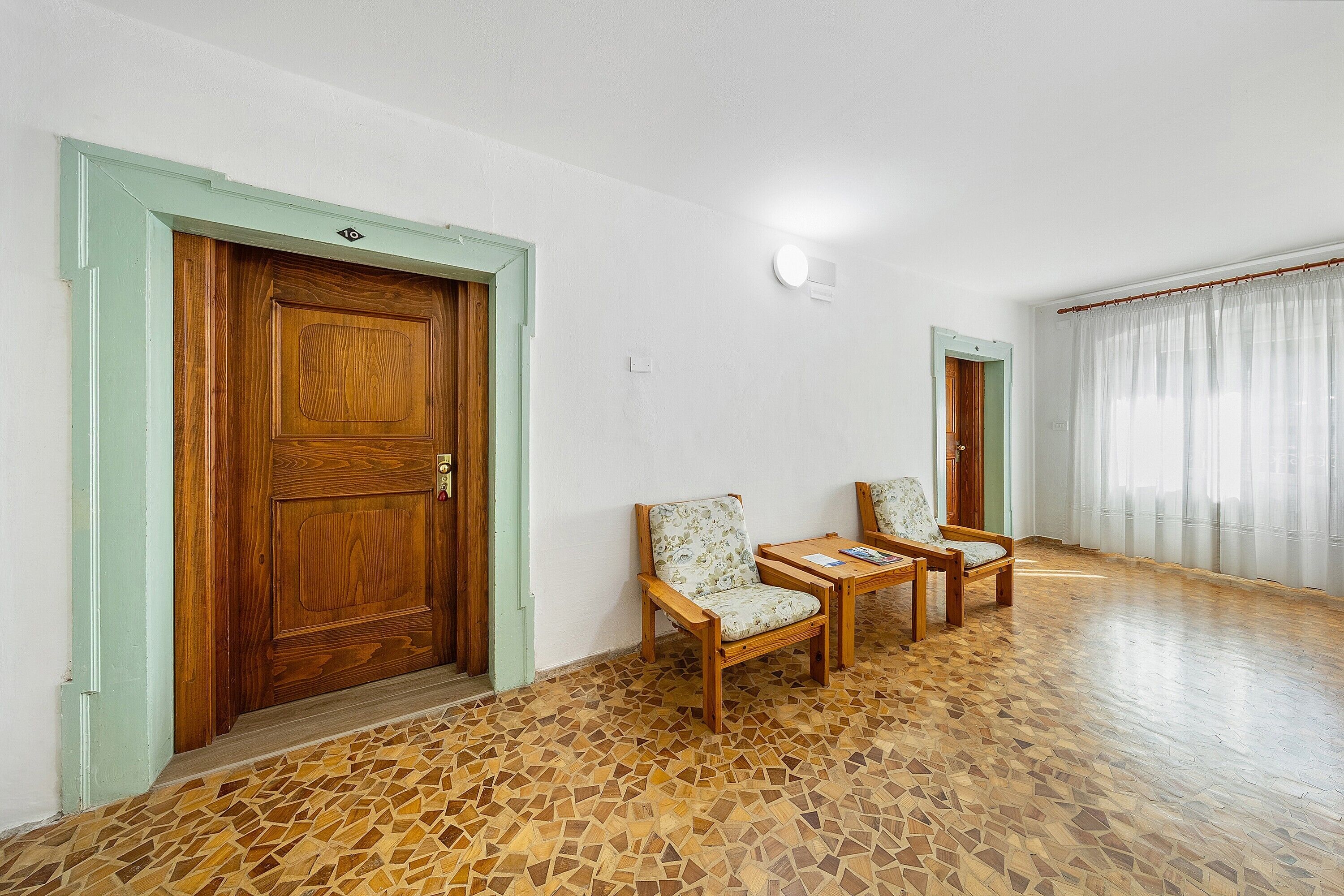 Una habitació amb una porta de fusta, dues cadires, una taula petita i un paviment amb dibuixos.