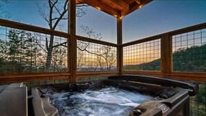 A hot tub with a view of a forested landscape through a wooden structure with glass panels. - Stunning Mtn Views | Hot Tub | Game Room | EV (Sevierville)
