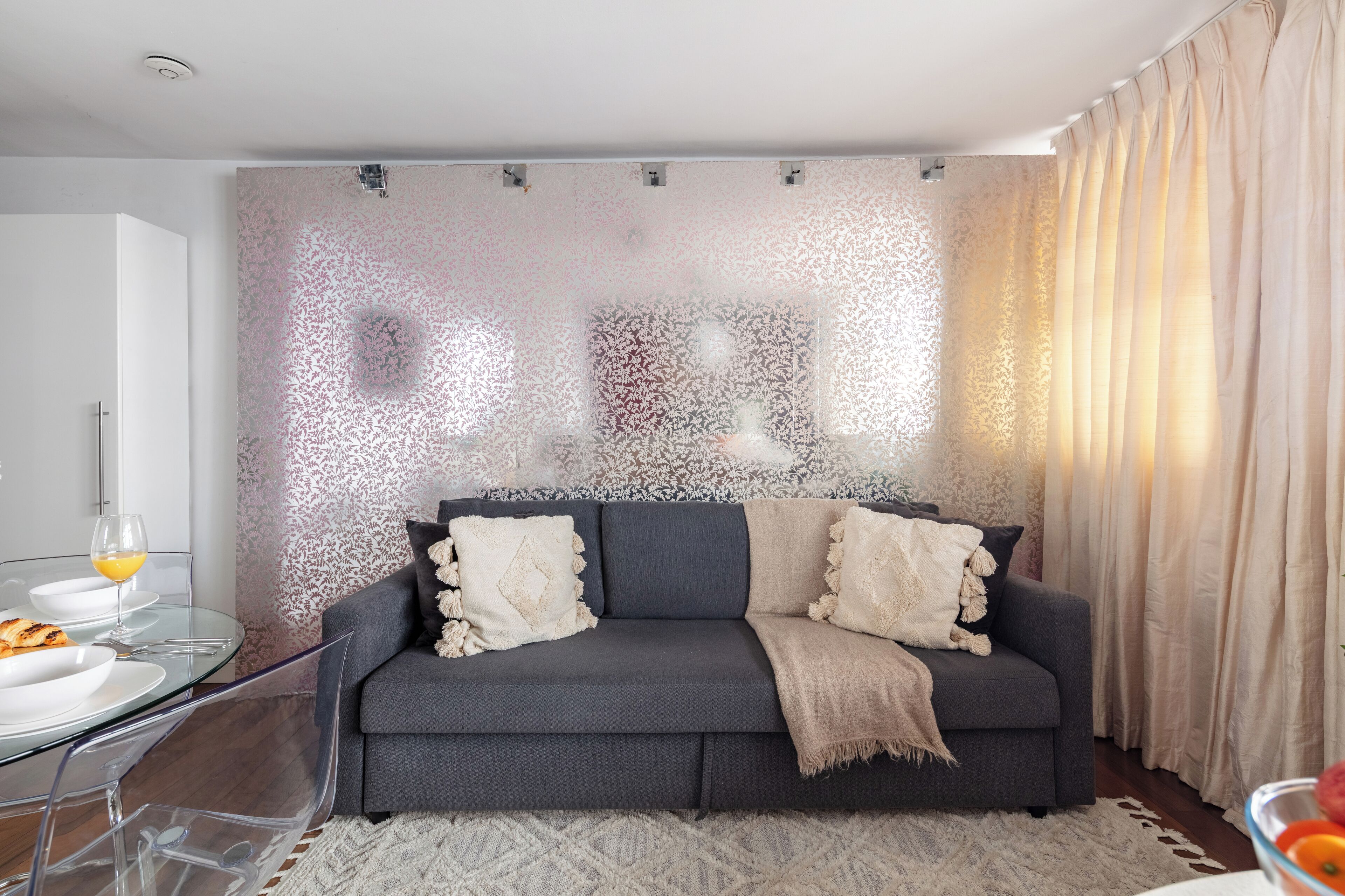 Ein modernes Wohnzimmer mit grauem Sofa, transparentem Glastisch und Wandpapier mit Muster.