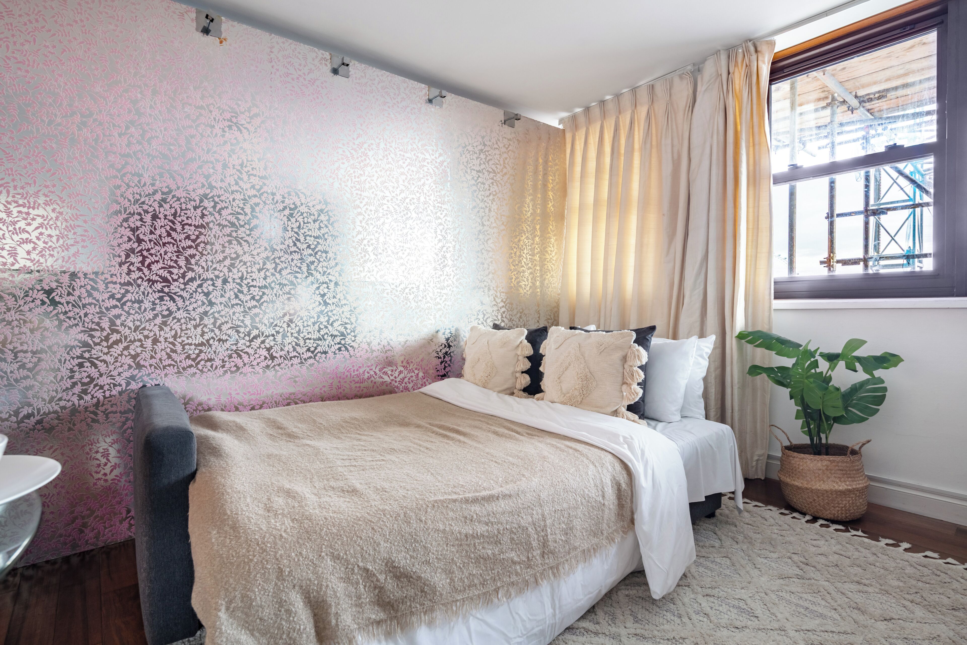 A bedroom with a bed, a window with curtains, a plant, and a patterned wall.