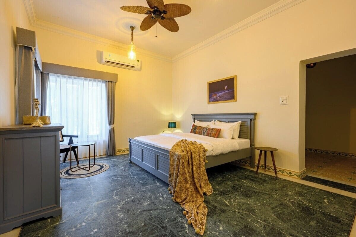 A bedroom with a bed, a desk, a chair, a ceiling fan, an air conditioner, and a window with curtains.