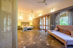 Quarto de hotel com cama, sofá, ventilador de teto e uma grande janela com cortinas.