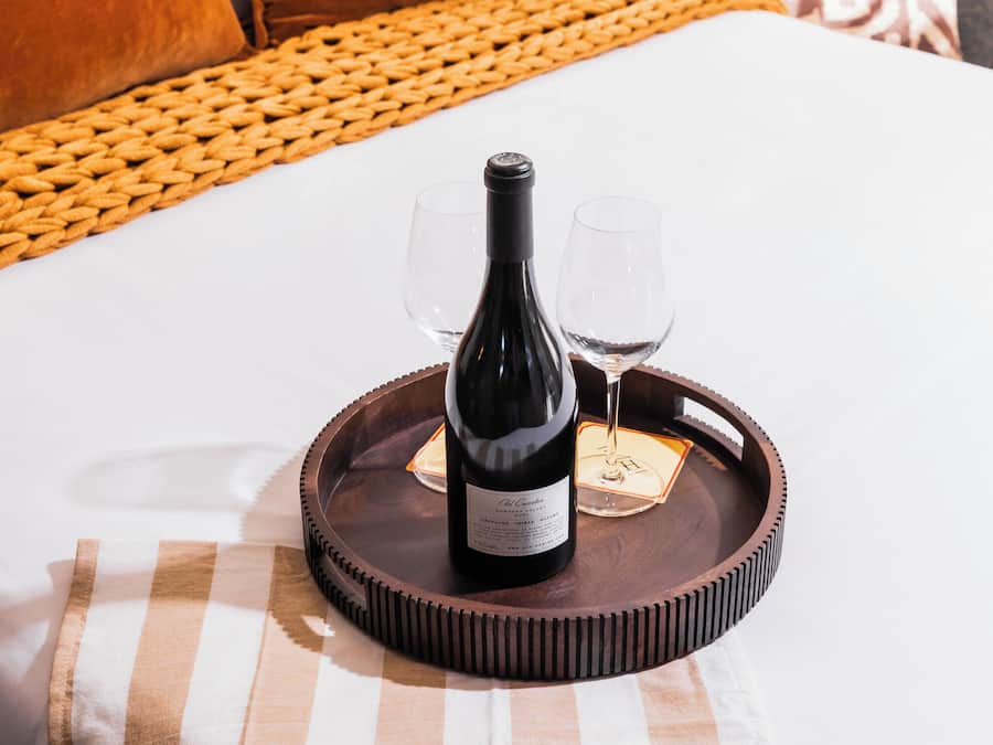 A bottle of wine on a tray with two glasses on a bed with a checkered bedspread.