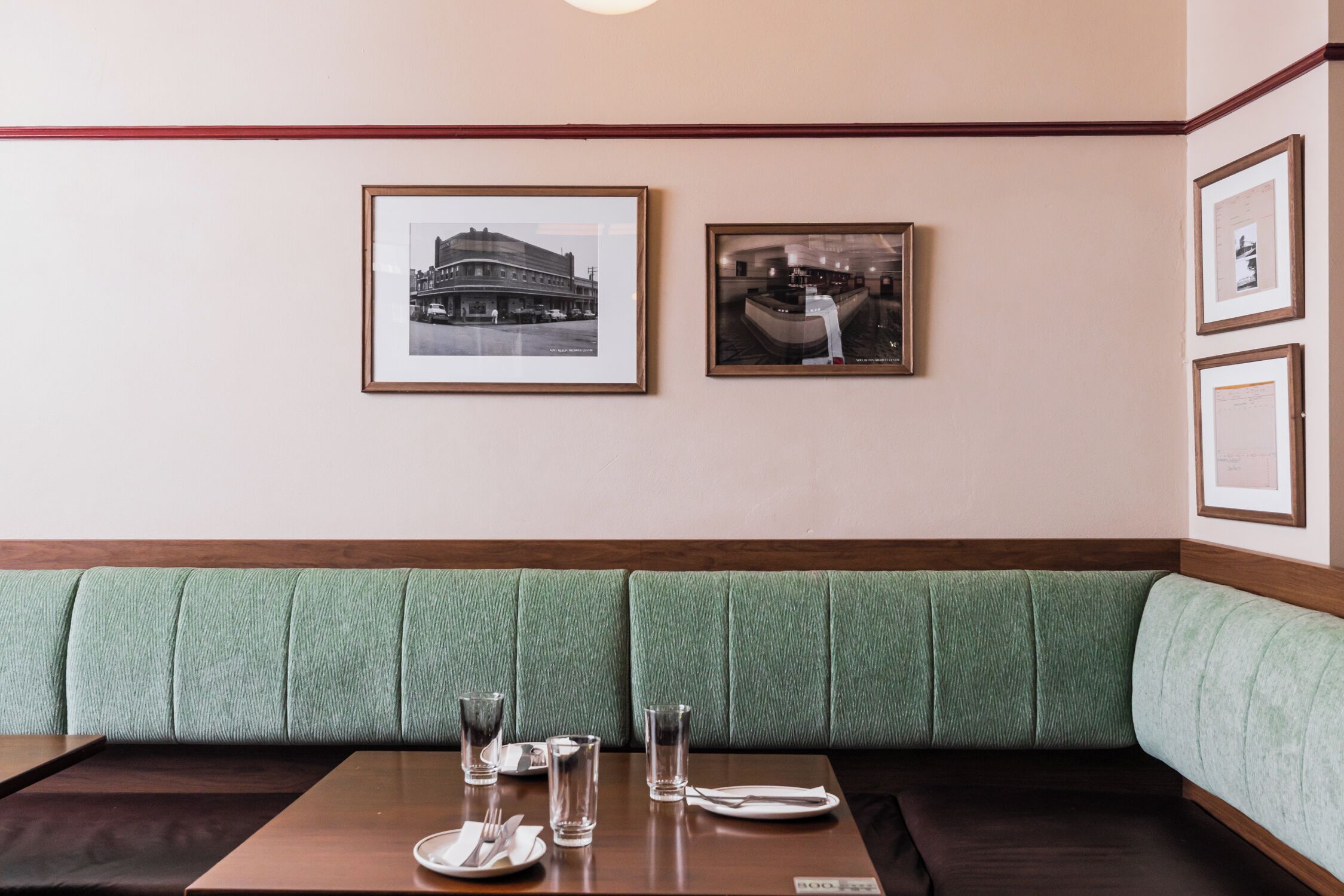 A restaurant with a green upholstered booth seating area, framed photographs on the wall, and a table set with plates, glasses, and cutlery.
