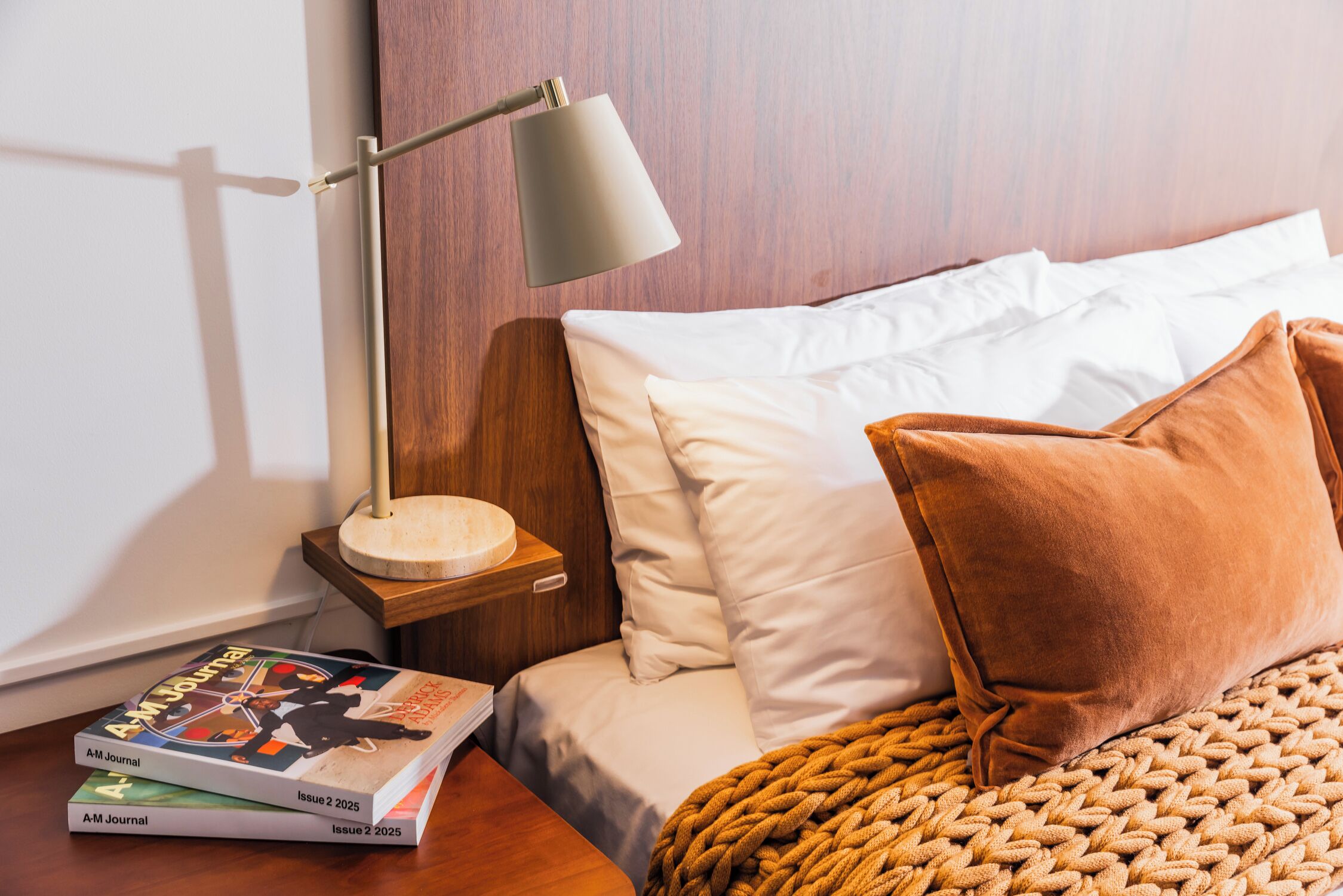 Una cama bien tendida con almohadas blancas y marrones, un cabecero de madera y una lámpara de noche. Hay dos libros sobre una mesita de noche de madera junto a la cama.