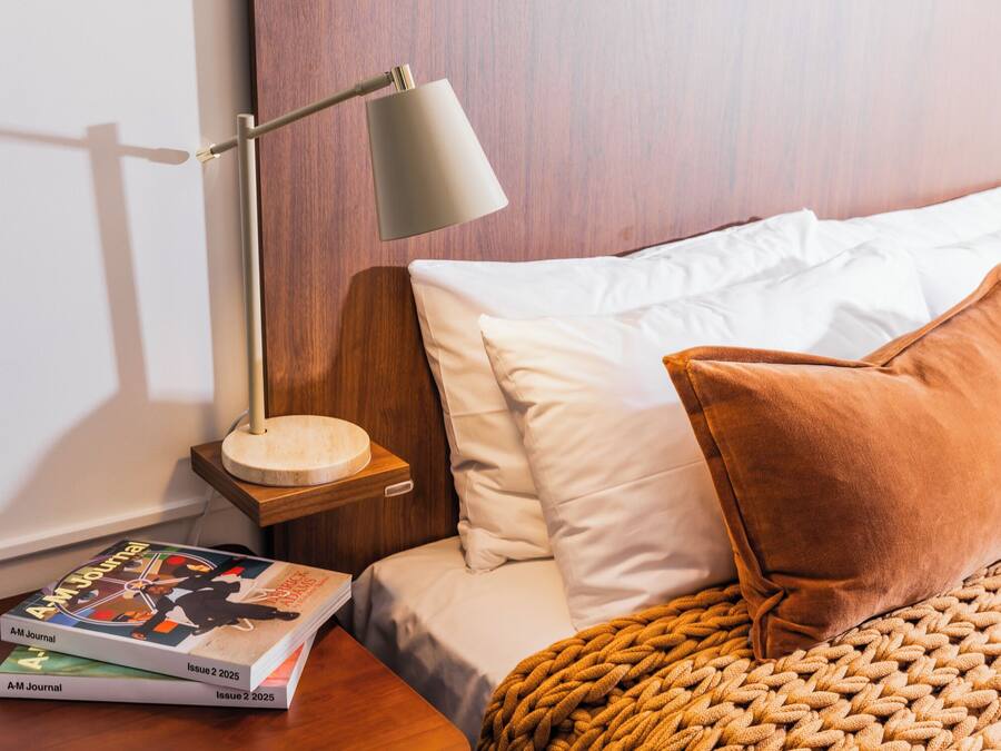Una cama bien tendida con almohadas blancas y marrones, un cabecero de madera y una lámpara de noche. Hay dos libros sobre una mesita de noche de madera junto a la cama.