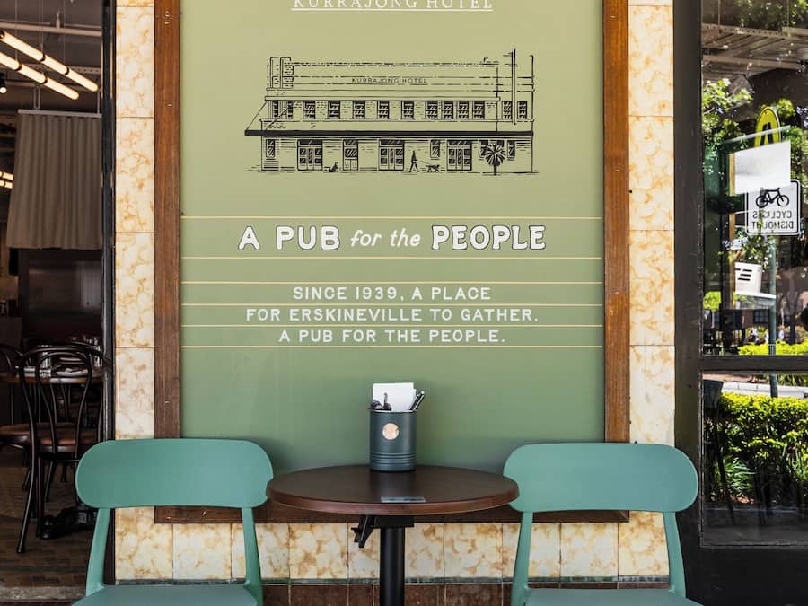 Un letrero en marco para el Hotel Kurrajong con un mensaje histórico, dos sillas verdes y una mesa al frente.