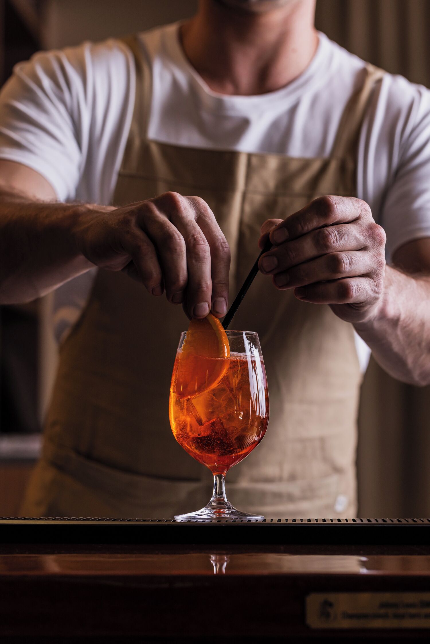 A person in a white shirt and apron preparing a drink with an orange slice and a straw in a glass.