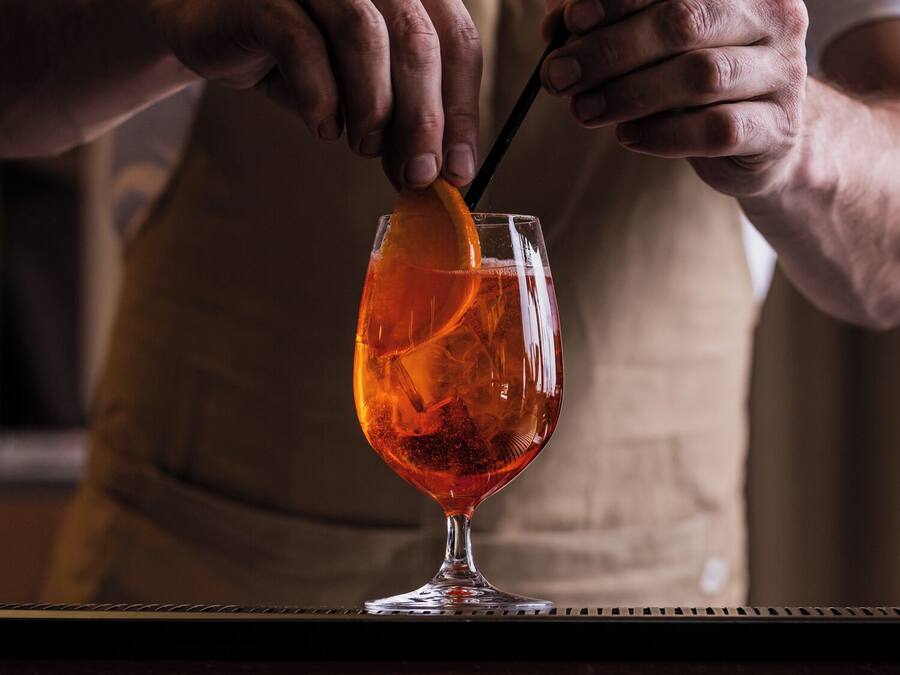 A person in a white shirt and apron preparing a drink with an orange slice and a straw in a glass.