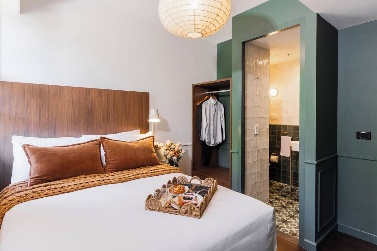 Un lit bien fait, avec un plateau contenant des articles pour le petit-déjeuner, une tête de lit en bois et un porte-manteau suspendu, dans une pièce dont un mur est peint dans une teinte verte, et où l’on aperçoit la salle de bain par une porte ouverte.