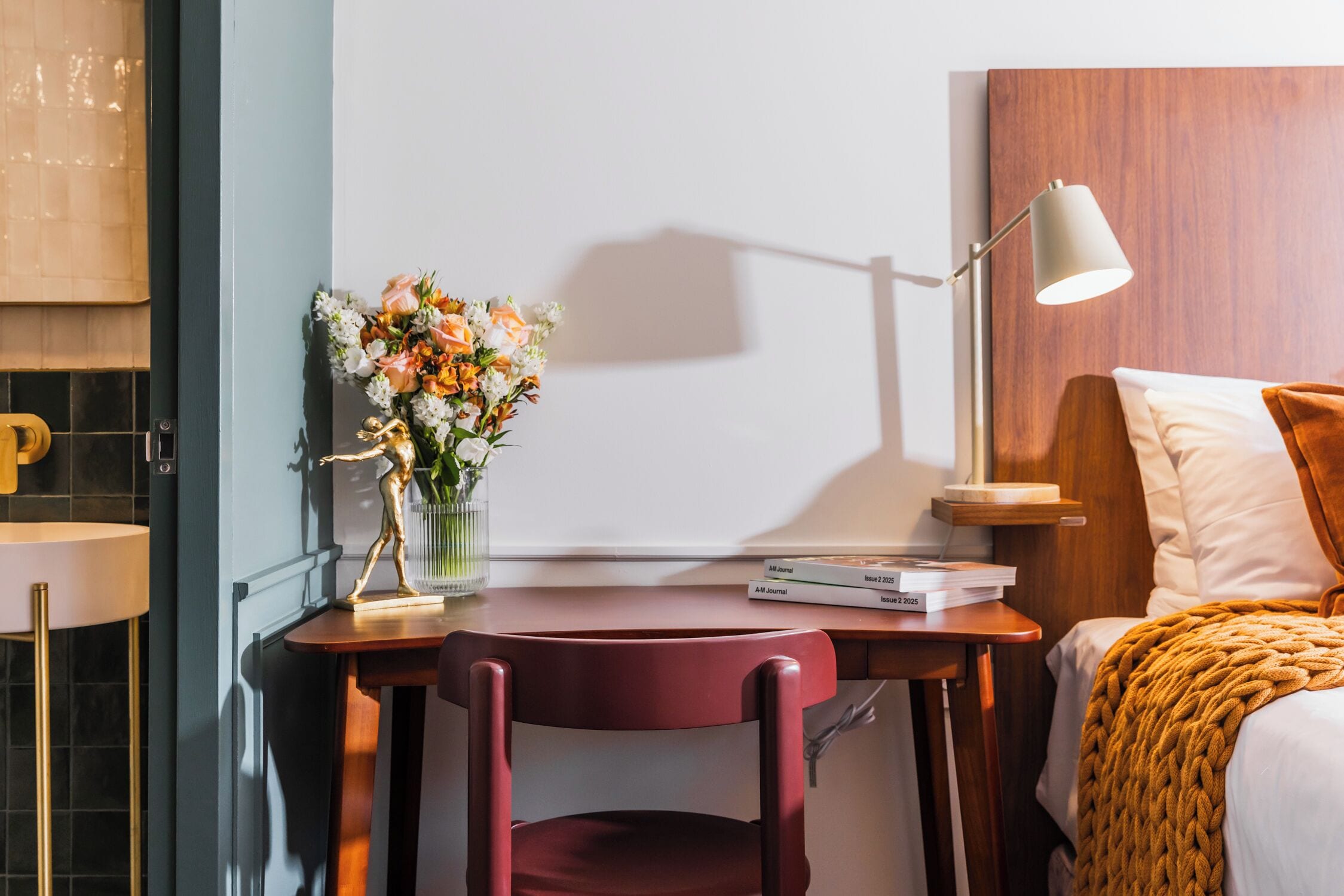 Ein Hotelzimmer mit Holz-Kopfteil, einem Schreibtisch mit Lampe, einem Stuhl, einem Bett mit Decke und einer Blumenvase.