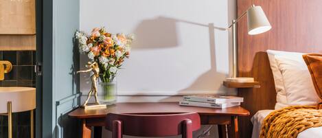 A hotel room with a wooden headboard, a desk with a lamp, a chair, a bed with a blanket, and a vase of flowers.
