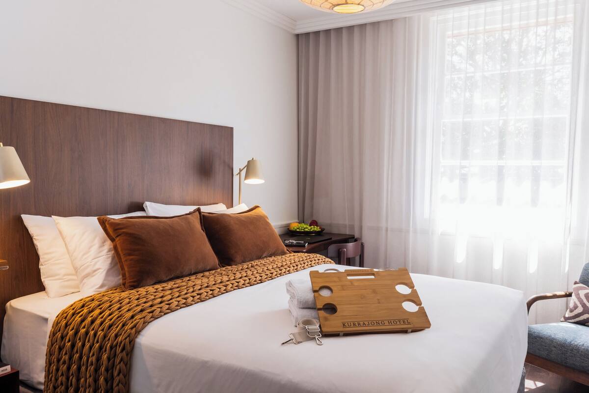 Un lit bien fait, recouvert d’une couverture marron, un plateau en bois avec une clé, et un oreiller orné du nom de l’hôtel.