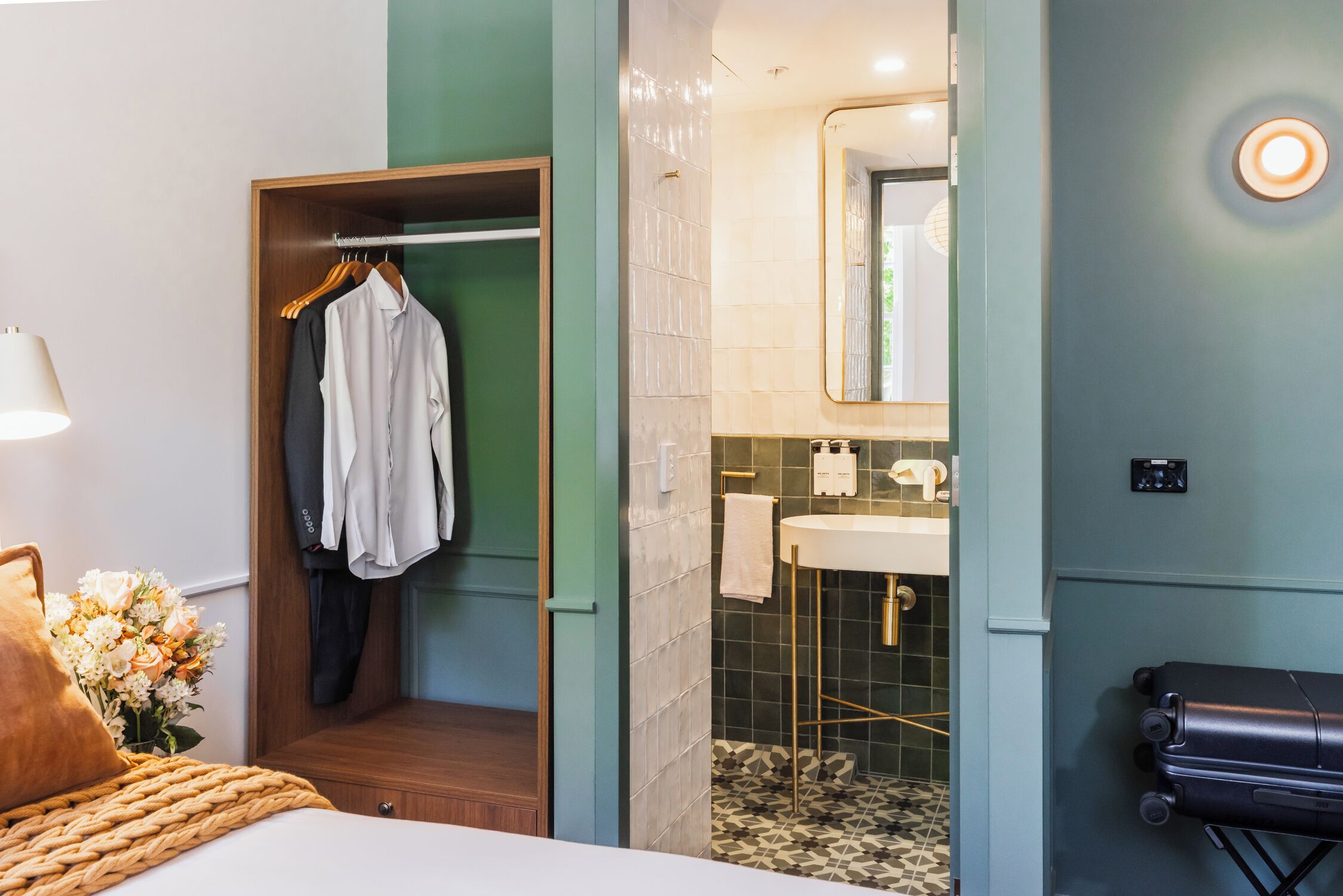 Una camera d'albergo con un letto, un armadio in legno, un bagno con lavandino e specchio, e una valigia sul pavimento.