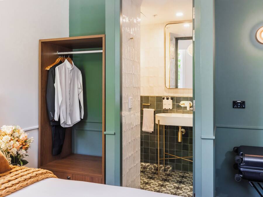 Ein Hotelzimmer mit einem Bett, einem Holzschrank, einem Badezimmer mit Waschbecken und Spiegel sowie einem Koffer auf dem Boden.
