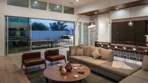 A spacious living area with a sofa, chairs, and a coffee table. There is a dining area with chairs and a table. The room has large windows and a kitchen area with cabinets. - Carbon @ Elemento49 - Pool & Spa - Sleeps 16 (Indio)
