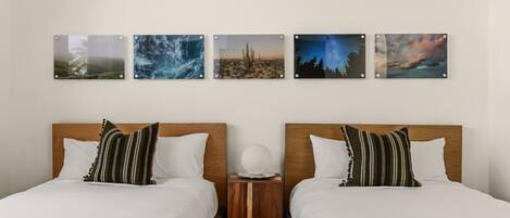 A hotel room with two beds, each with white bedding and striped pillows. There are four framed pictures on the wall above the beds.
