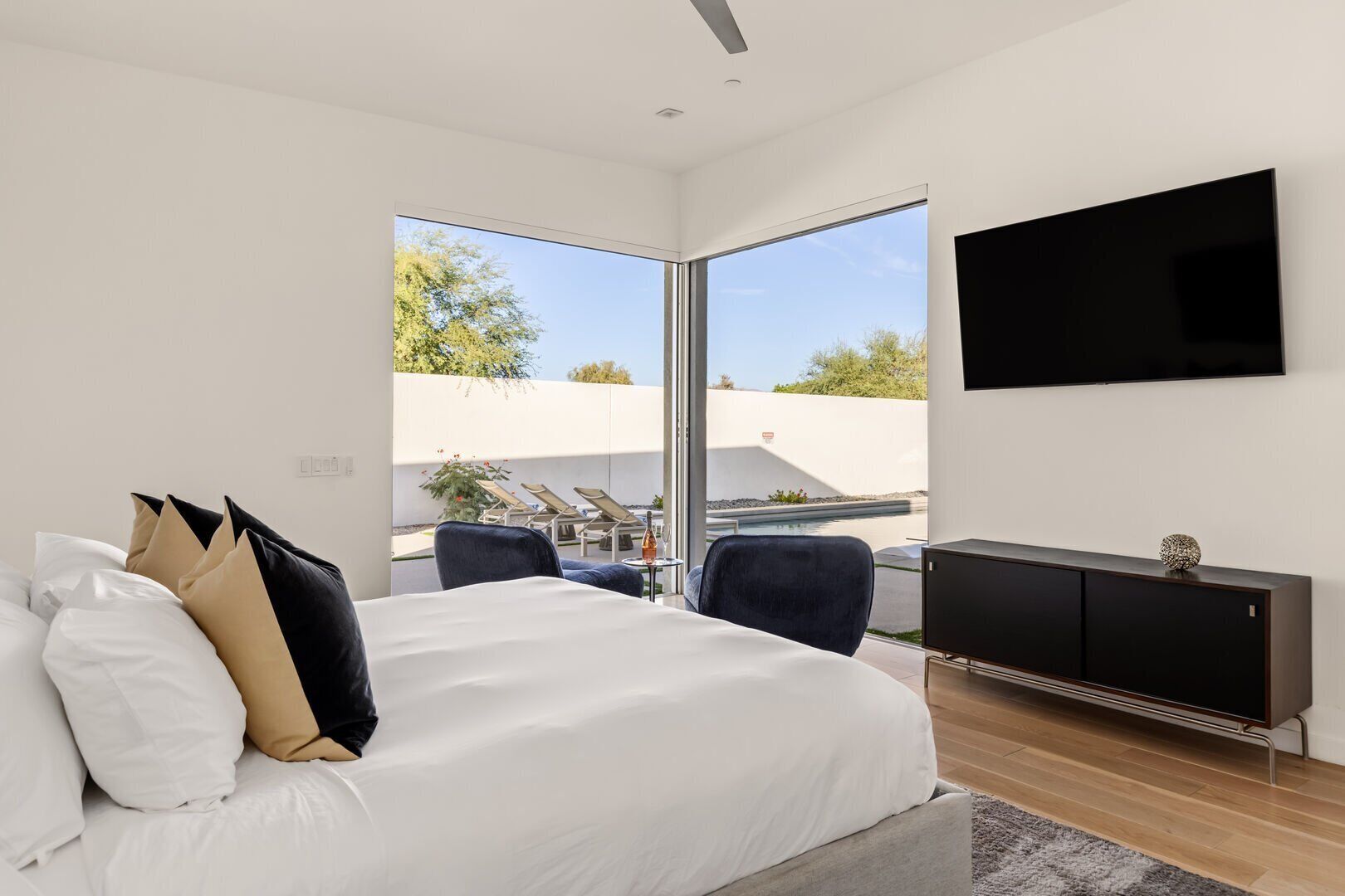 Une chambre moderne avec un grand lit, un téléviseur fixé au mur et une vue sur un espace extérieur aménagé avec des chaises et une piscine.