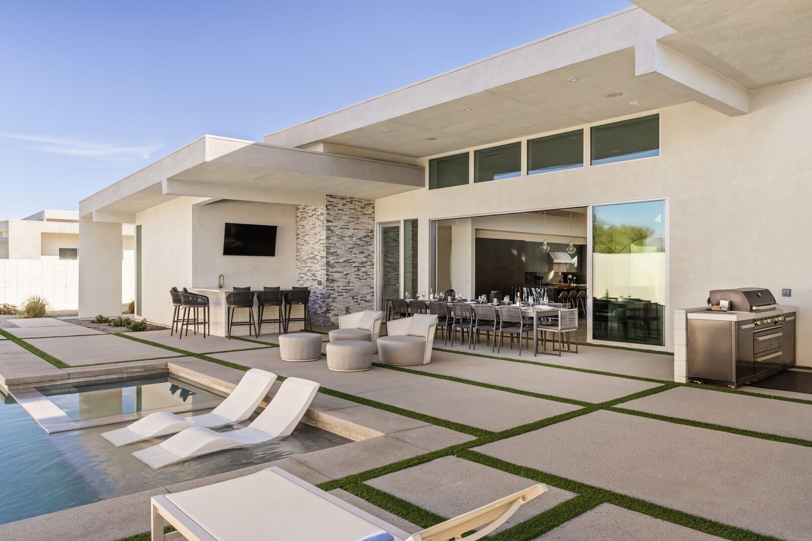 A modern outdoor patio with a pool, lounge chairs, a dining area, and a barbecue grill.