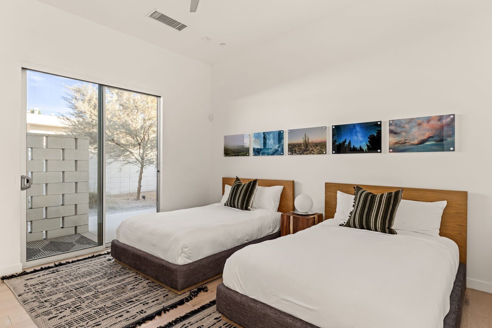 A modern hotel room with two beds, a sliding glass door to a balcony, and a series of landscape paintings on the wall.