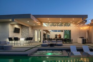 A modern outdoor living area with a pool, seating, and a kitchen visible through glass doors. - Mercury @ Elemento49 | Pool & Spa | Sleeps 16 (Indio)