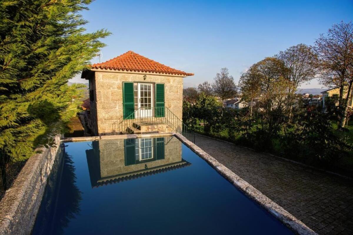 A stone building with a tiled roof and green shutters, adjacent to a clear swimming pool.
