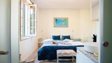 Quarto com cama, mesa de cabeceira, um banco, uma janela e um quadro na parede.
