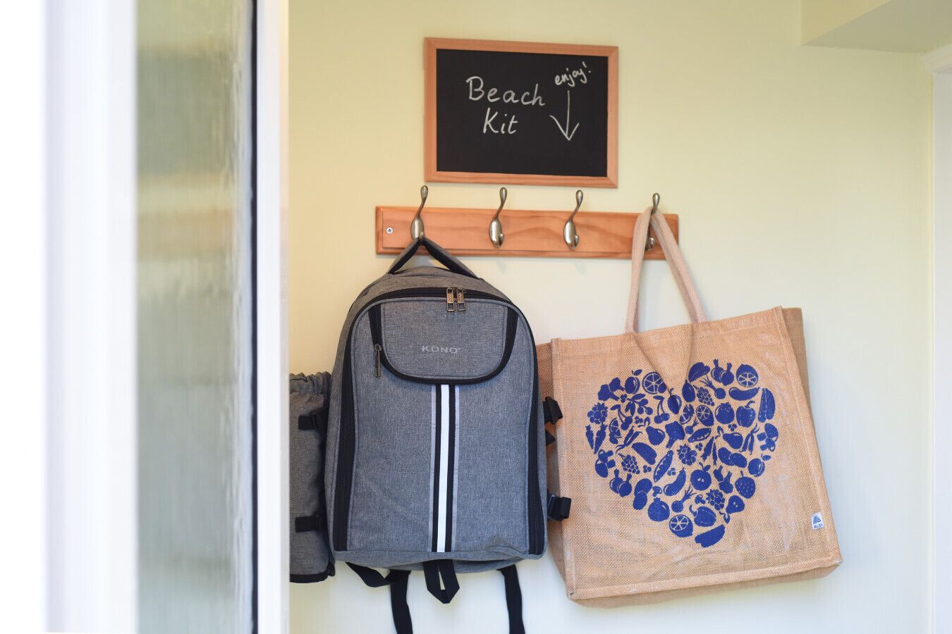Un crochet fixé au mur, sur lequel sont accrochés un sac à dos gris et un sac fourre-tout marron orné d’un motif en forme de cœur.