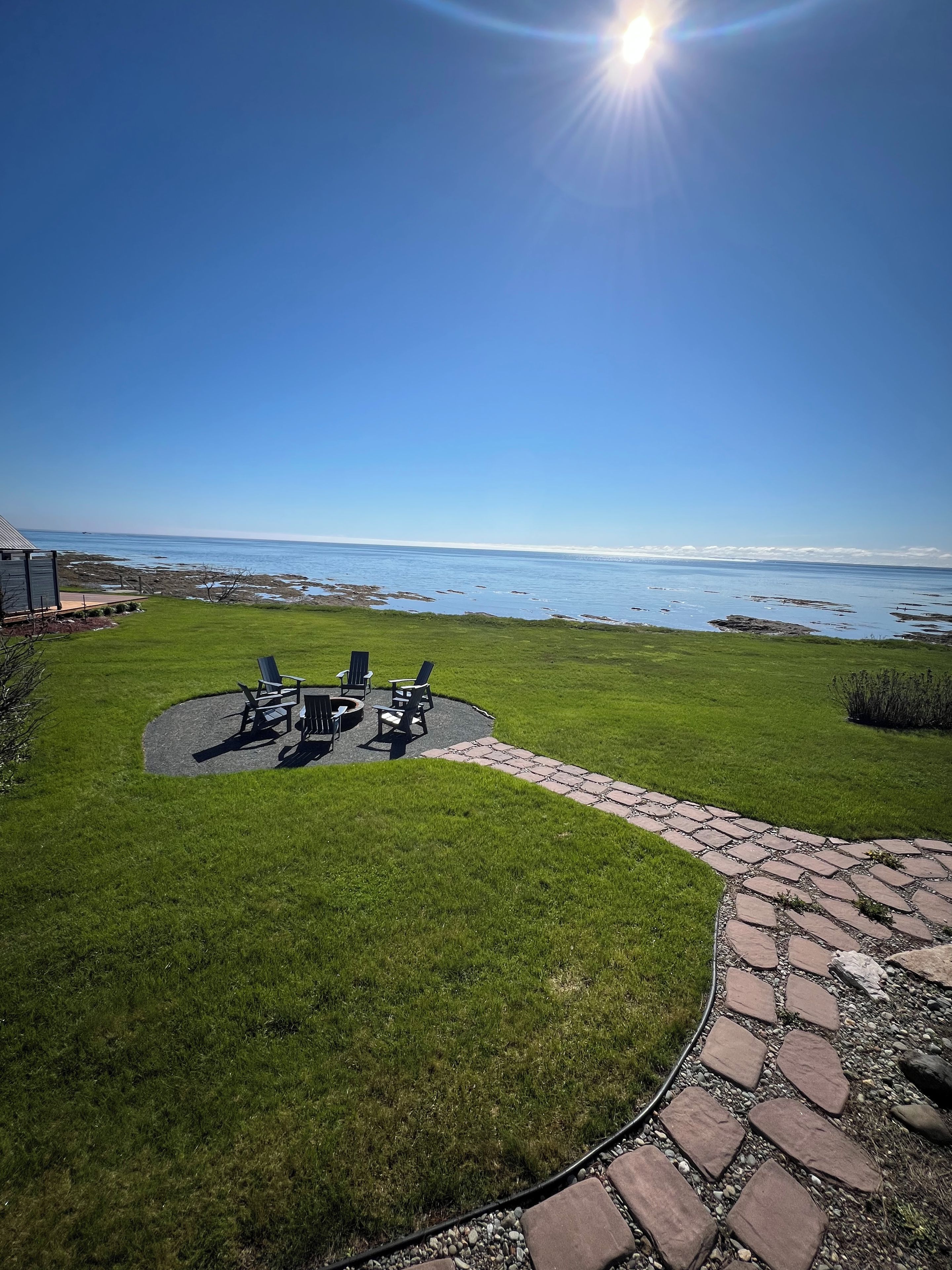 En terrasse med havemøbler og en stenkøbssti, der fører til et græsarbejde med udsigt over havet.