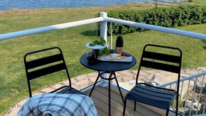 A deck with two chairs, a small table with a bottle of wine, grapes, and a bowl of fruit.