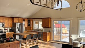 A modern kitchen with wooden cabinets, stainless steel appliances, a dining area with a table and chairs, and a view of the outside through large windows.