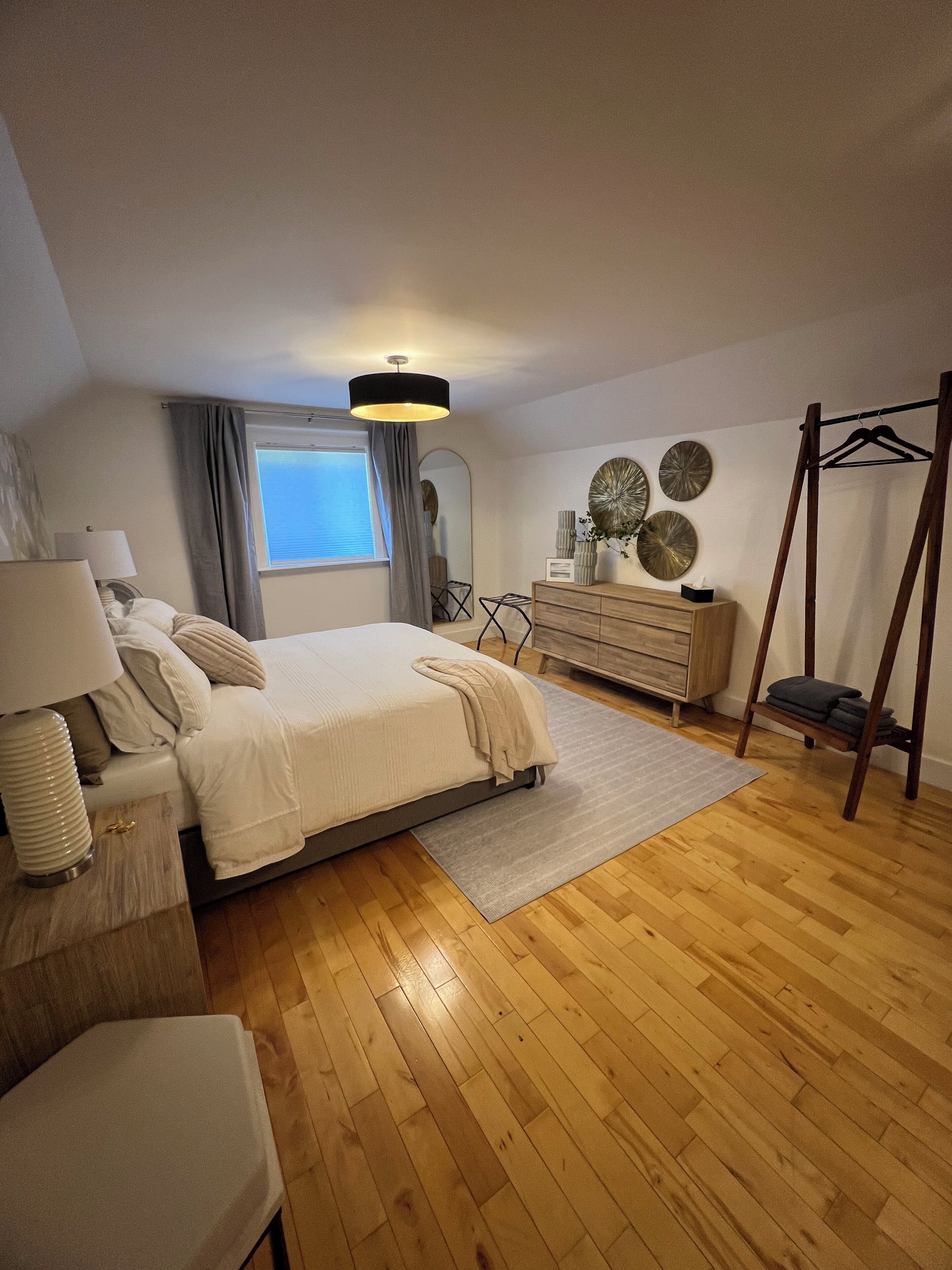 A bedroom with a bed, bedside table, lamp, dresser, mirror, and a window.