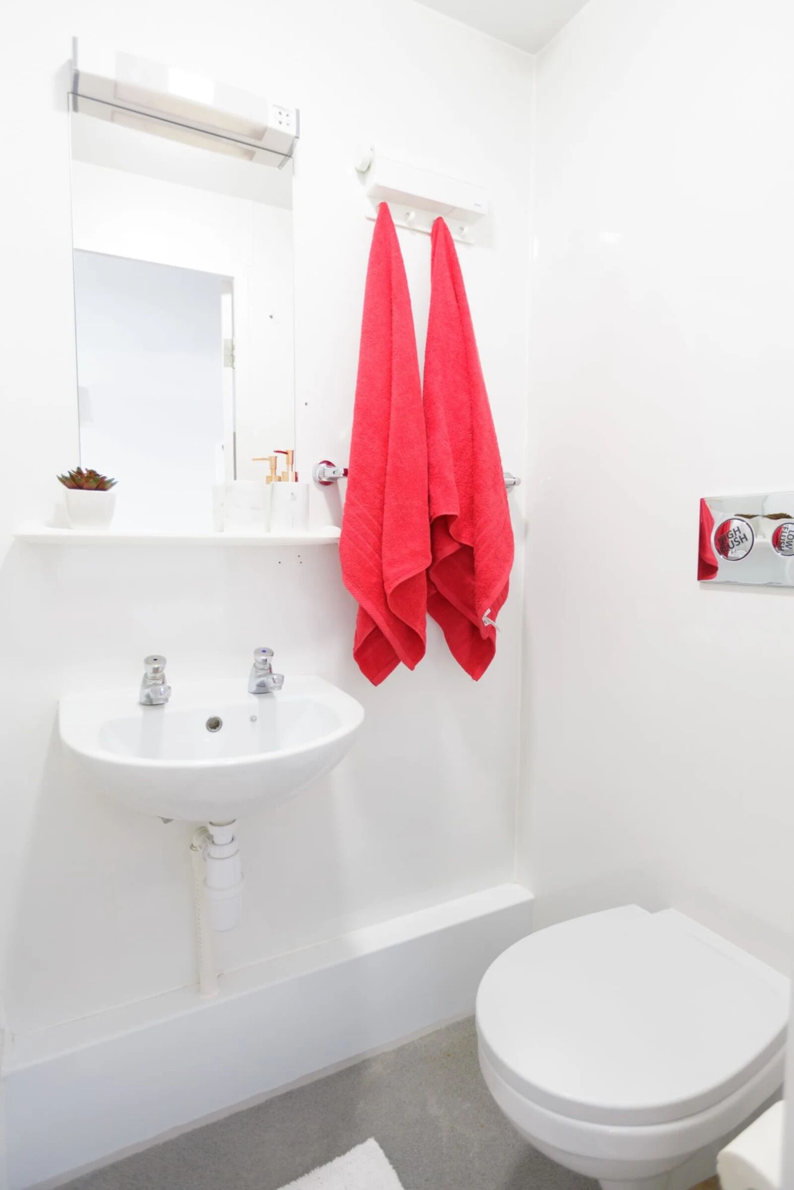 Une salle de bain avec un lavabo blanc, deux robinets, un miroir, une serviette rouge accrochée, des toilettes blanches et une baignoire blanche.