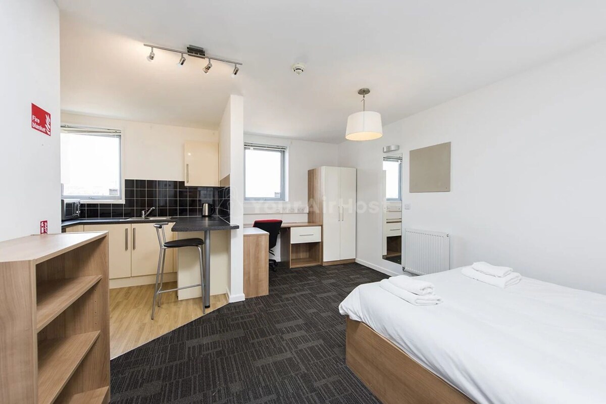 Quarto de hotel moderno com cama, escrivaninha, cadeira, cozinha compacta e banheiro.