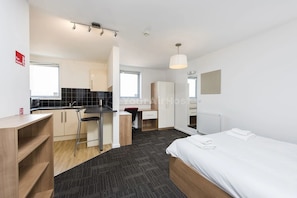 Quarto de hotel moderno com cama, escrivaninha, cadeira, cozinha compacta e banheiro.