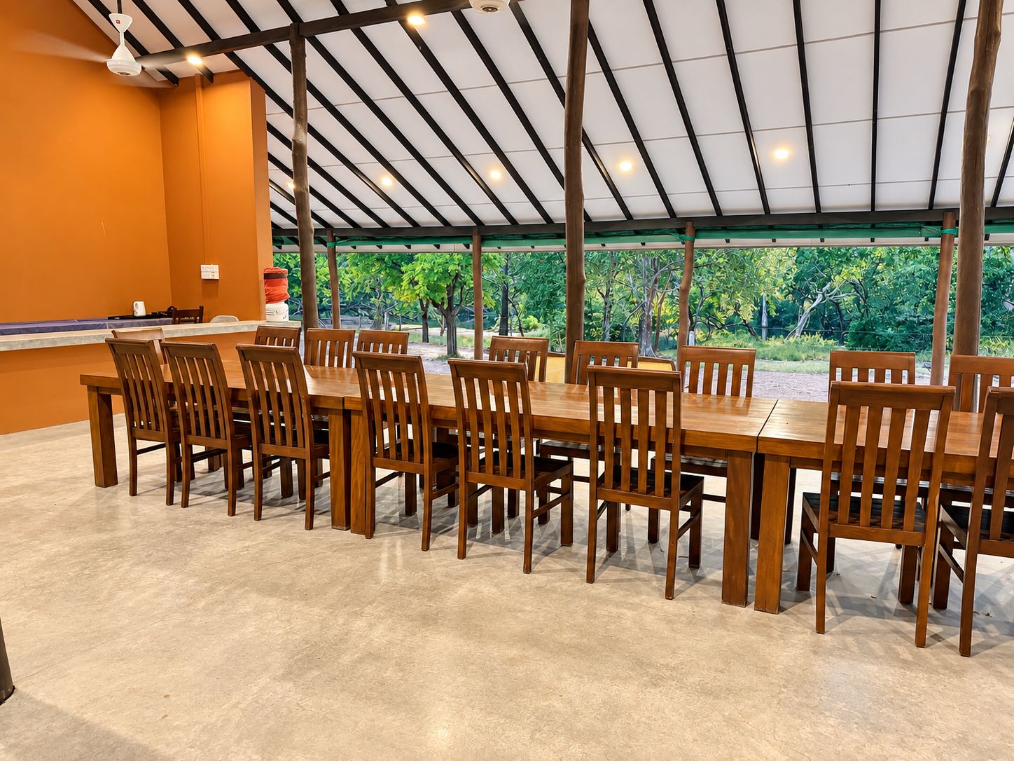 Un área de comedor techada con una mesa larga y sillas, un ventilador de techo y vista a la vegetación exterior.