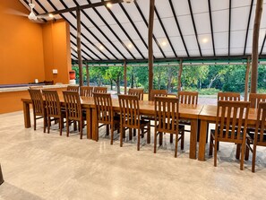 Een overdekt eetgedeelte met een lange tafel en stoelen, een plafondventilator en uitzicht op groen.