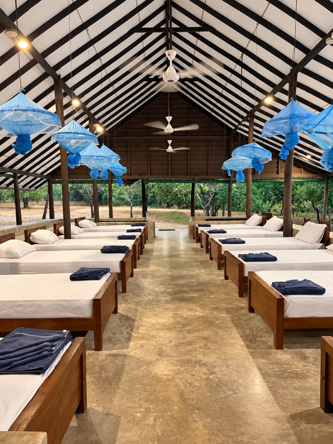 A row of beds with white linens and blue towels, under a high ceiling with a fan and blue umbrellas.
