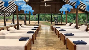 A row of beds with white linens and blue towels, under a high ceiling with a fan and blue umbrellas.