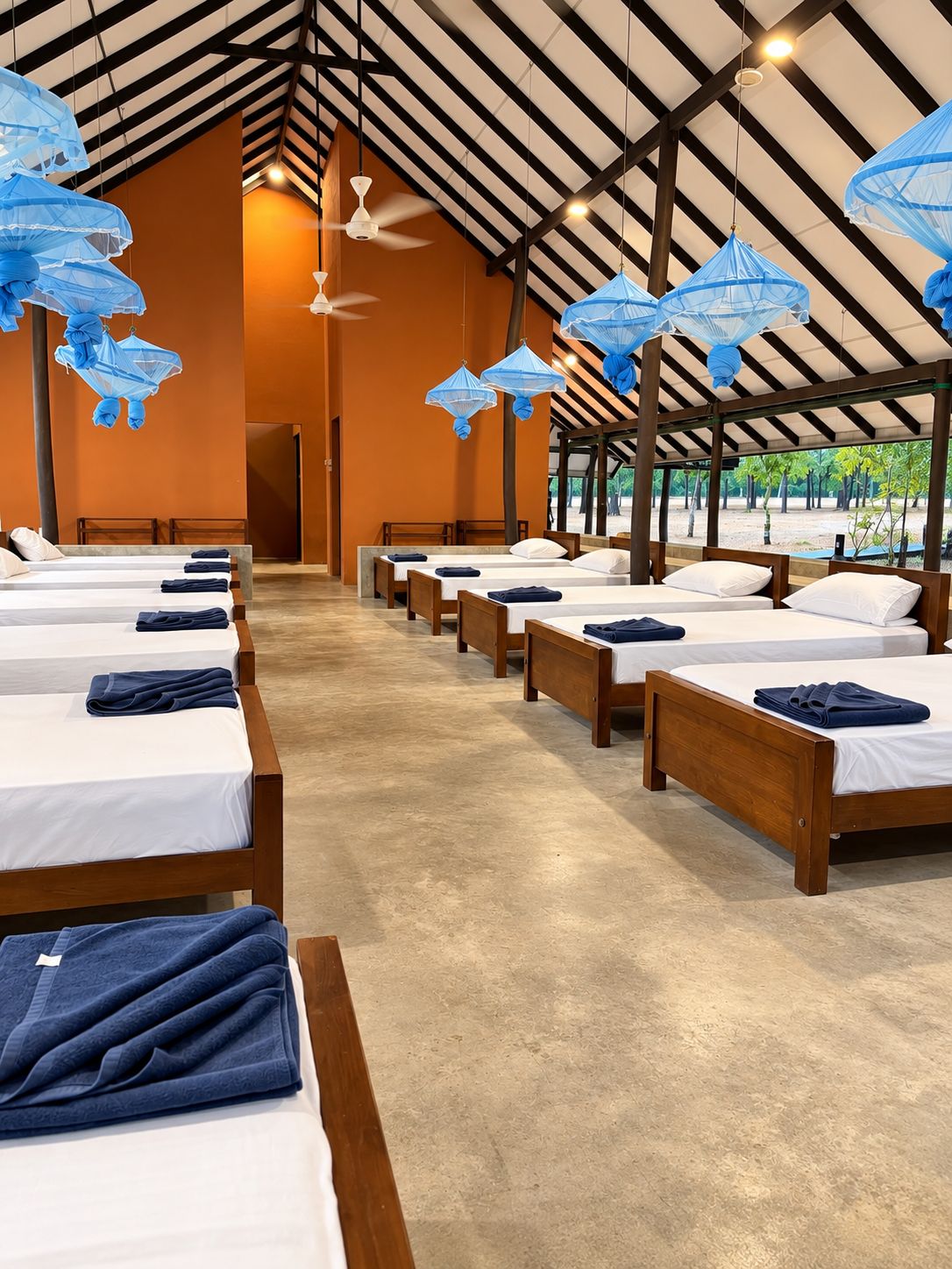Habitación de hotel con varias camas dispuestas en filas, techo alto con vigas a la vista y ventiladores de techo.