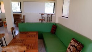 Un divano verde con un cuscino decorato, un tavolo di legno con sedie e una lavagna in una stanza con pavimento piastrellato.
