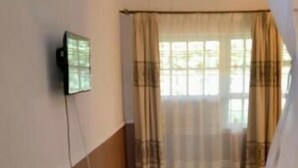 A room with a bed, a television mounted on the wall, and a window with curtains. - Trotters Hotel (Kabati)