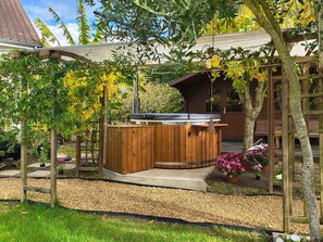 Un bain à remous extérieur en bois, surmonté d’une pergola et entouré de gravier et de verdure.