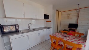 Une cuisine compacte avec des meubles blancs, un four à micro-ondes et un évier. On y trouve un coin repas avec une table et des chaises, ainsi qu’un téléviseur dans un coin.