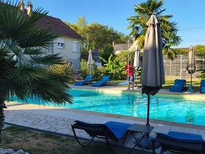 Una piscina con sillones de descanso y sombrillas, rodeada de vegetación y una casa.
