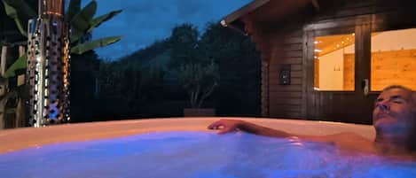 Una persona relajándose en un jacuzzi con una cabaña de madera al fondo.