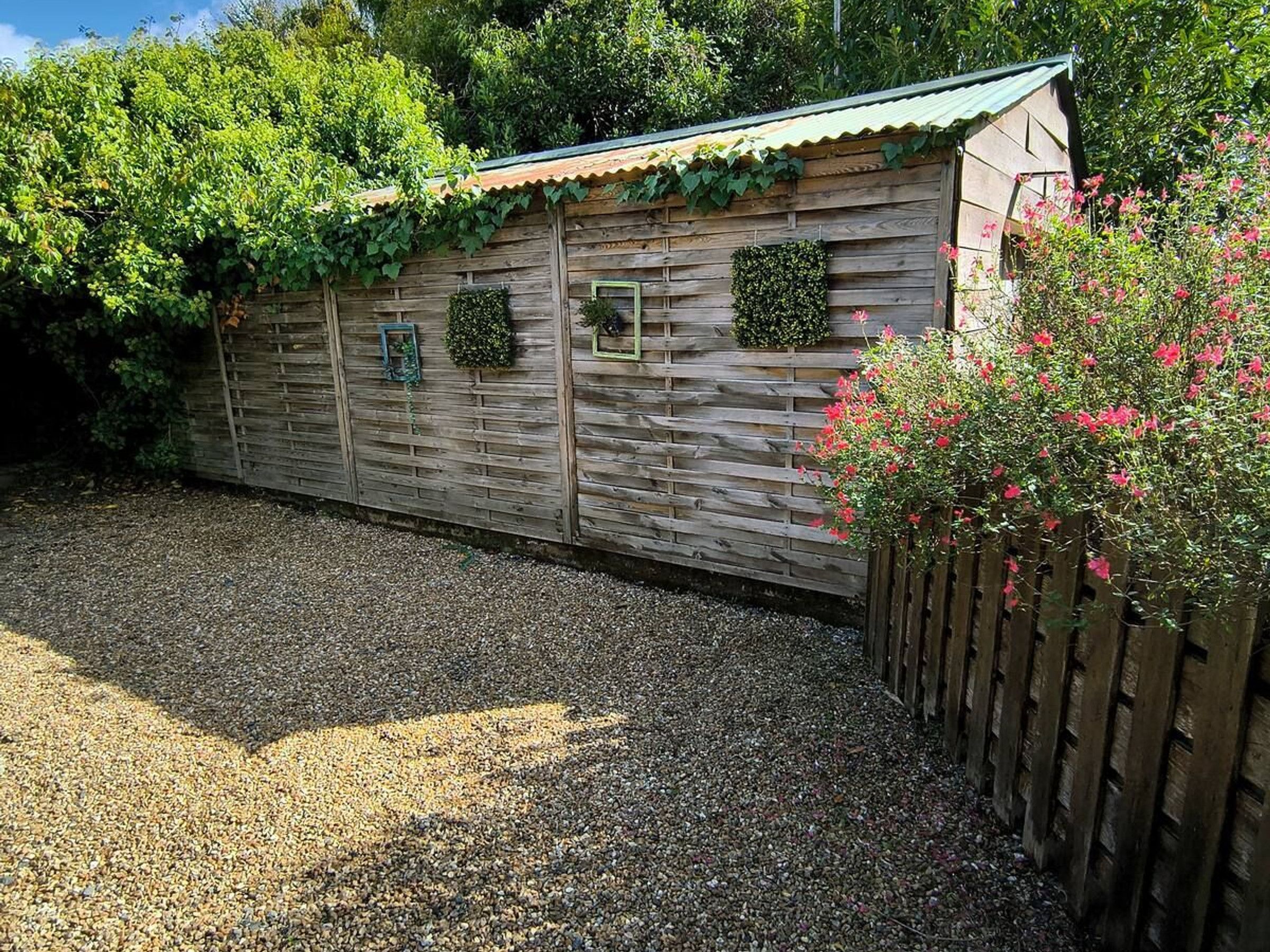 A wooden shed with a corrugated metal roof, surrounded by gravel and greenery.