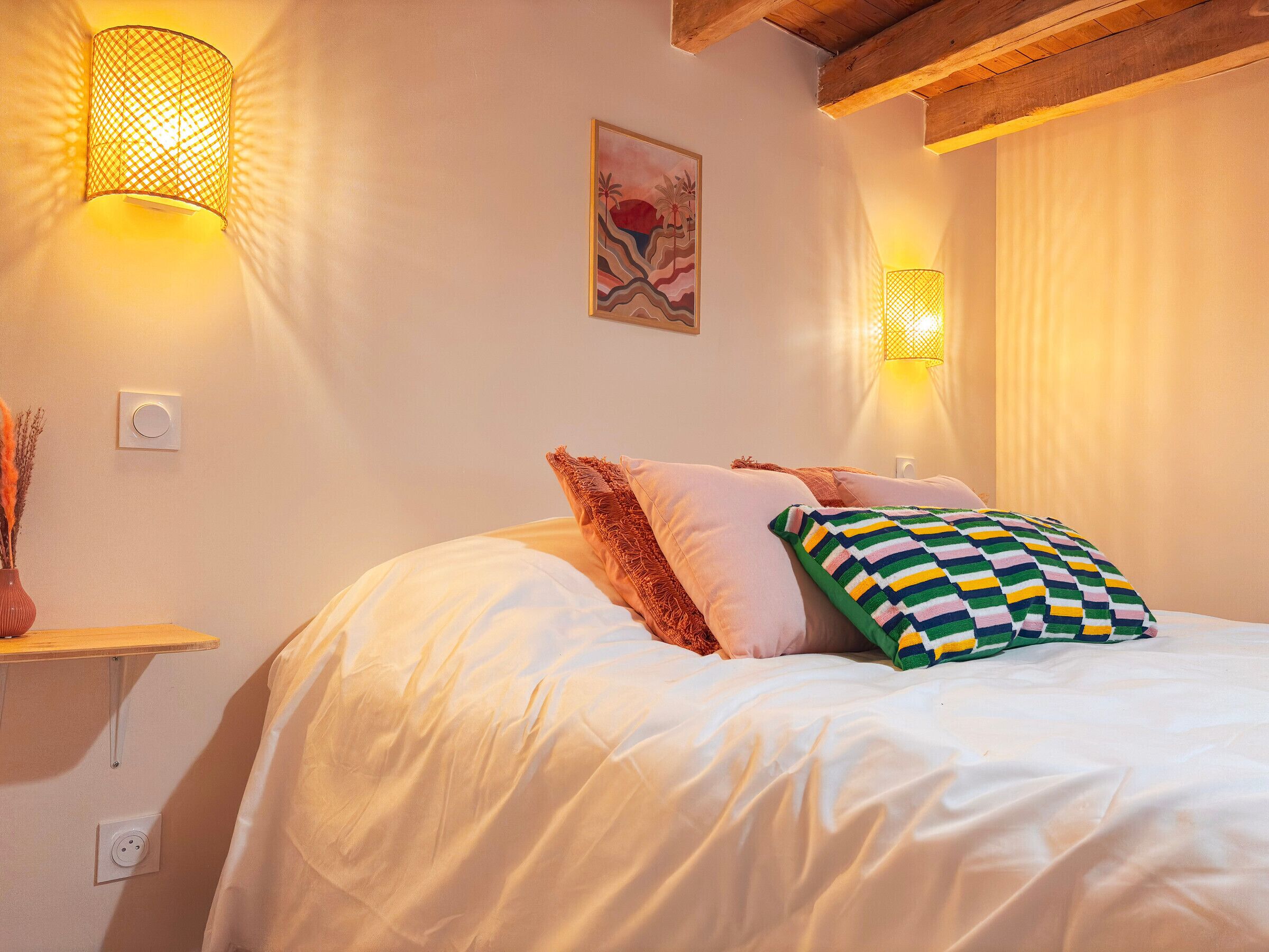 A neatly made bed with white bedding and colorful pillows, two wall-mounted lamps, and a painting on the wall.